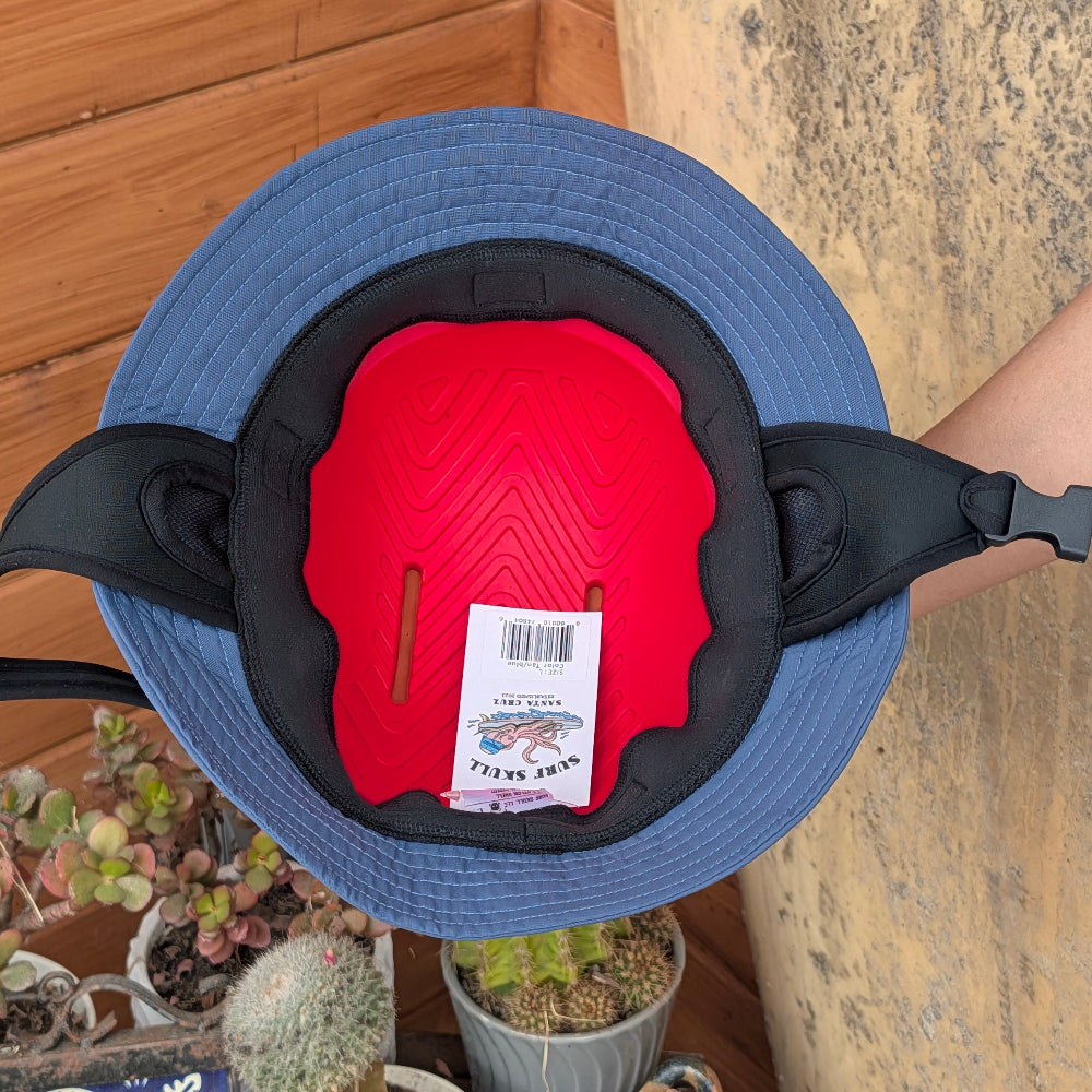A blue wide-brim sun hat with a black adjustable strap and a bright red inner foam insert, shown from above on a wooden surface with potted plants in the background.